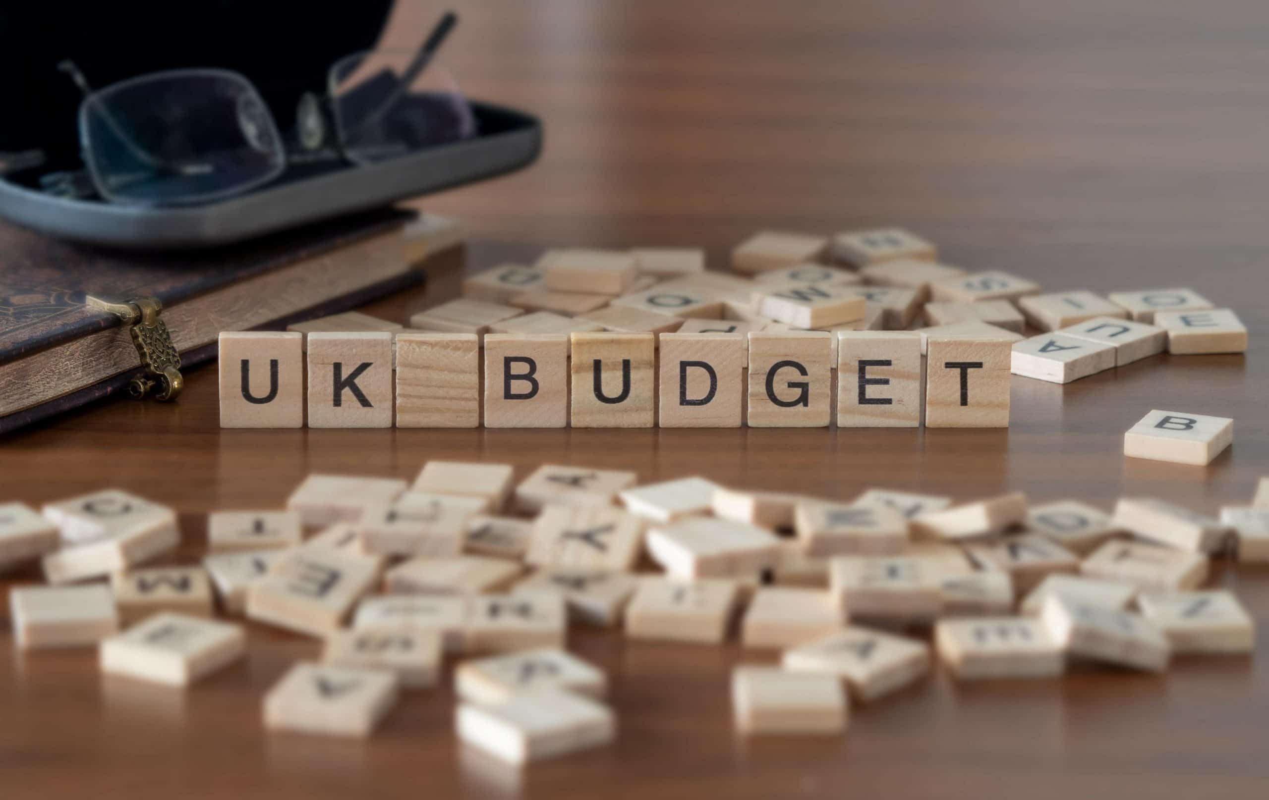 uk budget word represented by wooden letter tiles on a wooden table with glasses and a book